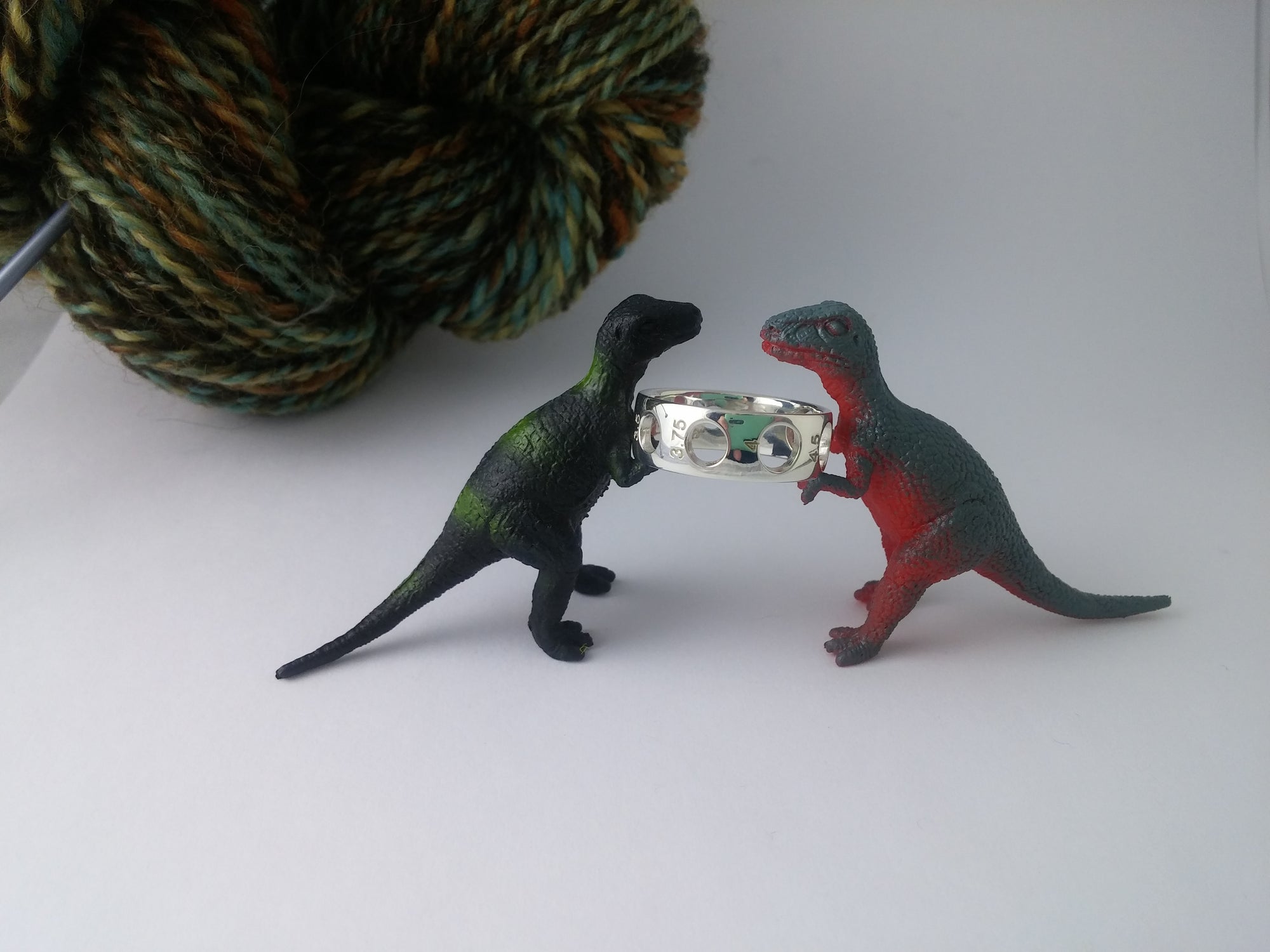 
          
            two tiny dinosaur toys holding a silver knitting needle gauge ring in front of handspun yarn
          
        